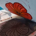 Linoleum block with a flower carved into it. A print of the flower, which is orange, is being pulled up. on February 4, 2026